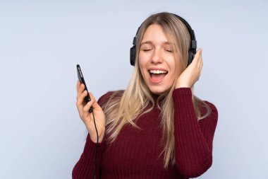 Soyutlanmış mavi arka planda genç sarışın kadın cep telefonuyla müzik dinliyor ve şarkı söylüyor.