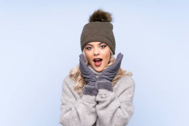 Kışlık şapkalı genç kız izole edilmiş mavi arka planda ve yüzünde sürpriz bir ifade var.