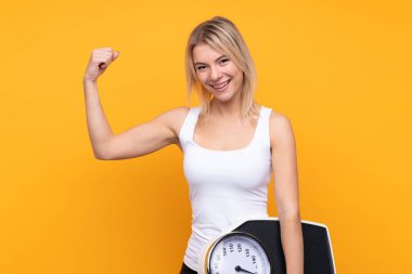 Sarışın genç bir Rus kadın izole edilmiş sarı arka planda elinde bir tartı makinesiyle güçlü bir hareket yapıyor.