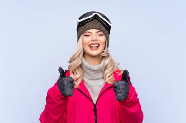 Snowboard gözlüklü genç kız izole edilmiş mavi arka planda başparmak hareketi yapıyor.