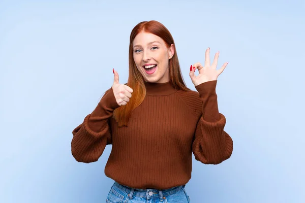 年轻的红头发女人在孤立的蓝色背景上表现出良好的体征和大拇指向上的姿势 — 图库照片