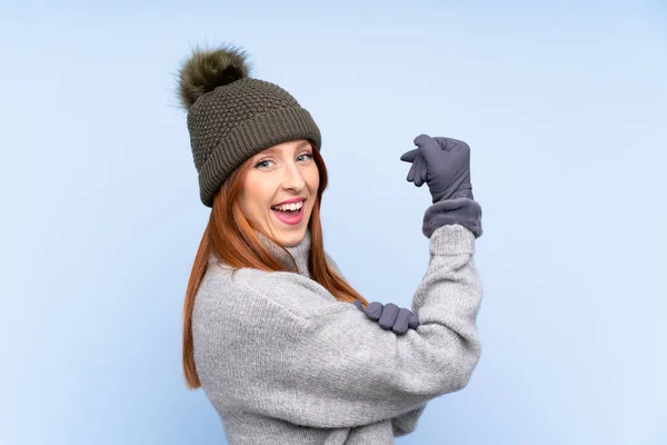 Young redhead Russian woman with winter hat over isolated blue background making strong gesture