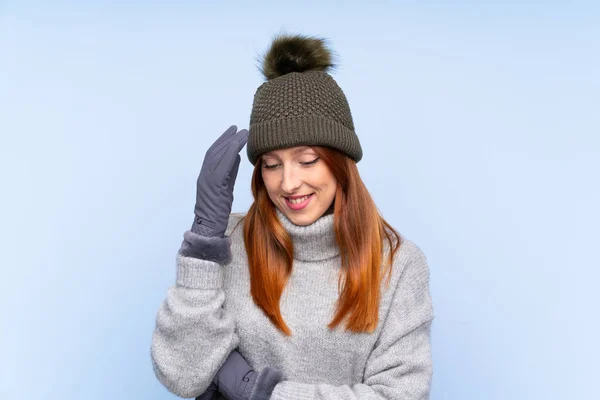 Young redhead Russian woman with winter hat over isolated blue background laughing