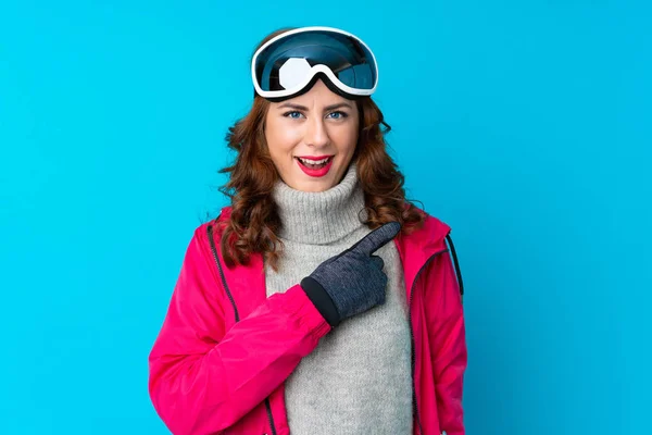 Lyžařka Snowboardovými Brýlemi Přes Izolovanou Modrou Stěnu Ukazující Prstem Stranu — Stock fotografie