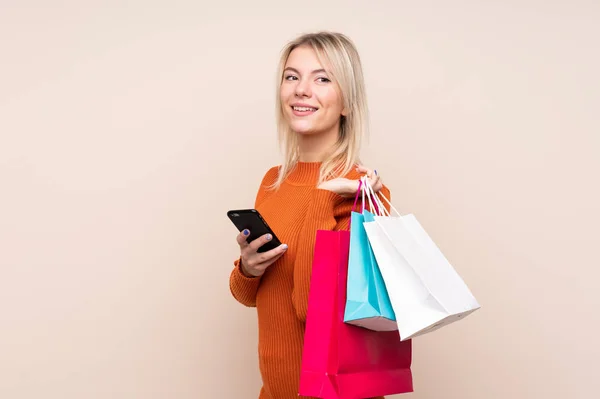 Ung Blond Kvinna Över Isolerad Bakgrund Håller Shoppingväskor Och Mobiltelefon — Stockfoto