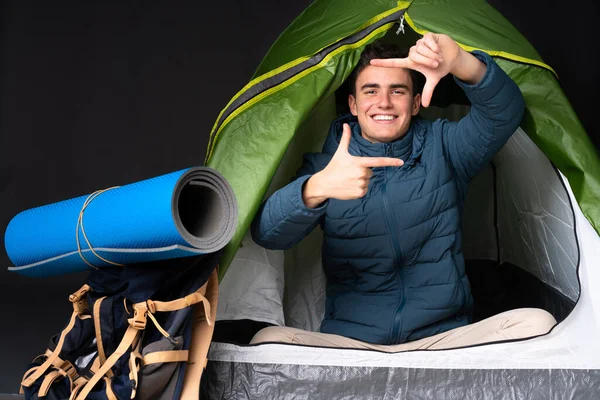 Teenager caucasian man inside a camping green tent isolated on black background focusing face. Framing symbol
