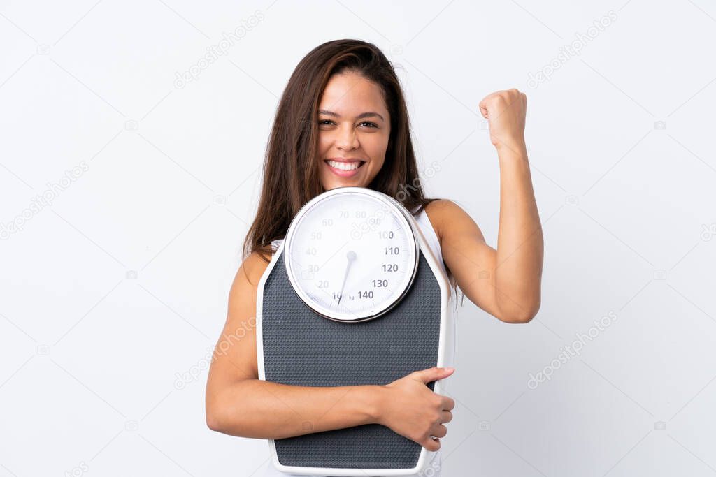 Young Brazilian girl holding a scale over isolated background with weighing machine and doing victory gesture