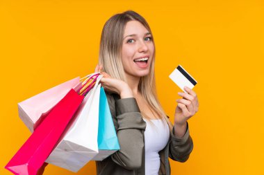 Sarışın, izole edilmiş, sarı arka planda genç bir kadın elinde alışveriş çantaları ve bir kredi kartı tutuyor.