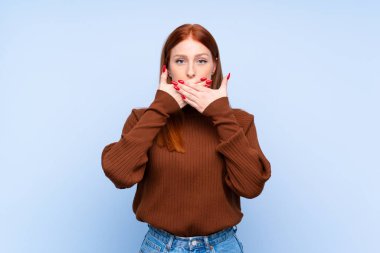 İzole edilmiş mavi arka planı olan genç kızıl saçlı kadın elleriyle ağzını kapatıyor.