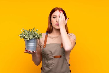 Genç, kızıl saçlı bahçıvan kadın, sarı arka planda bir bitkiyi elinde tutarak bir gözü kapatıyor.