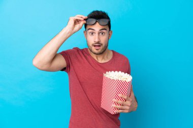 Soyutlanmış mavi arka planı olan yakışıklı bir adam 3D gözlüklü ve elinde koca bir kova patlamış mısırla şaşırdı.