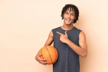 Genç Afrikalı Amerikalı adam bej arka planda izole edilmiş basketbol oynuyor ve yan tarafına işaret ediyor.