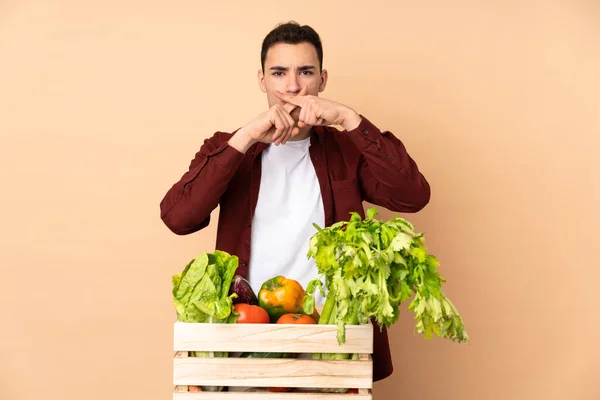 Farmer with freshly picked vegetables in a box isolated on beige background showing a sign of silence gesture