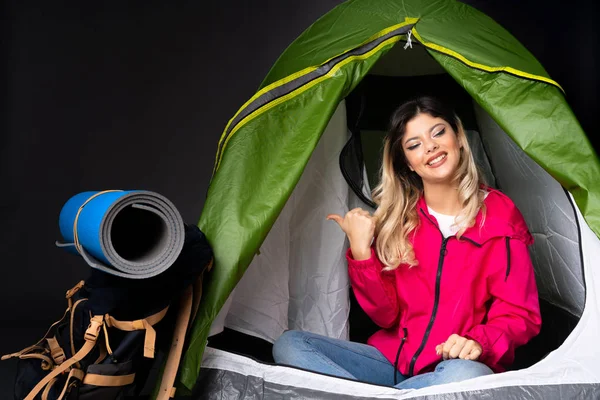 Teenager girl inside a camping green tent isolated on black background pointing to the side to present a product