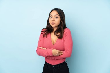 Genç melez kadın mavi arka planda izole edilmiş. Omuzlarını kaldırırken şüpheyle hareket ediyor.