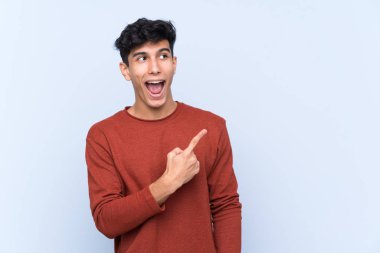 Arjantinli genç adam soyutlanmış mavi arka plan şaşırmış ve işaret ediyor.