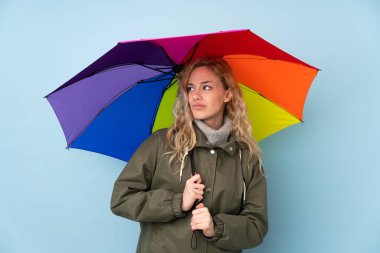 Mavi arka planda, üzgün bir ifadeyle izole edilmiş şemsiyeyi tutan genç sarışın kadın.