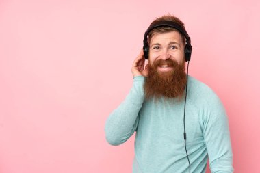 Uzun sakallı, soyutlanmış pembe arka planda müzik dinleyen kızıl saçlı adam.