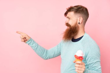 Kızıl saçlı, uzun sakallı, elinde kornet dondurması olan izole edilmiş pembe arka planı olan bir adam bir ürünü sunmak için kenarı işaret ediyor.