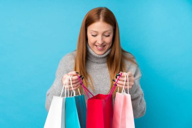 Genç kızıl saçlı kadın izole edilmiş mavi arka planda elinde alışveriş torbalarıyla içeriye bakıyor.