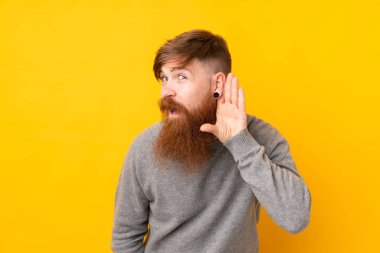 İzole edilmiş sarı arka planda uzun sakallı kızıl saçlı adam kulağına elini sürerek bir şeyler dinliyor.