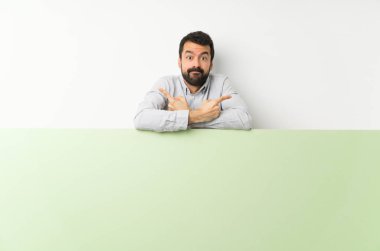 Sakallı genç yakışıklı adam, elinde kocaman boş bir pankart var. Yan taraftakileri işaret ediyor.