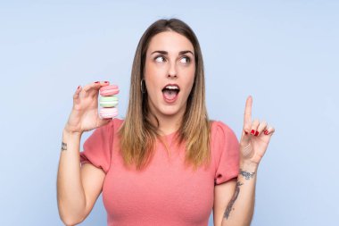 Young blonde woman over isolated blue background holding colorful French macarons and intending to realizes the solution