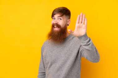 Uzun sakallı kızıl saçlı adam izole edilmiş sarı arka planda mutlu bir ifadeyle selamlıyor.