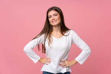 Soyutlanmış pembe arka planda genç bir kadın kolları kalçasında ve gülümsüyor.