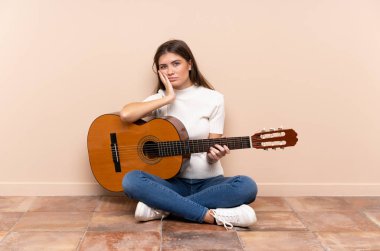 Gitarlı genç bir kadın mutsuz ve sinirli bir şekilde yerde oturuyor.