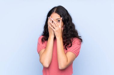 İzole edilmiş mavi arka planda süveter giyen, gözleri kapayan ve parmaklarından bakan melez bir kadın.