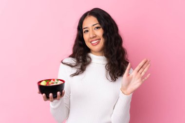 Bir kase erişteyi soyutlanmış pembe arka planda tutan melez kadın mutlu bir ifadeyle selamlıyor.