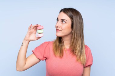 Soyutlanmış mavi arka planda genç sarışın kadın elinde renkli Fransız kurabiyeleriyle yukarı bakıyor ve gülümsüyor.