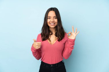 Mavi arka planda izole edilmiş genç melez kadın işaret ve baş parmak işareti gösteriyor.