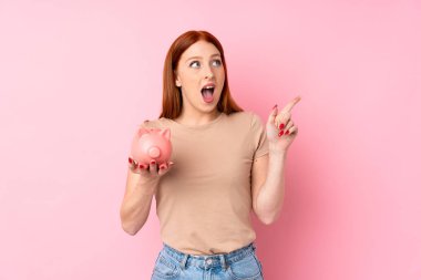 Soyutlanmış pembe arka planı olan genç kızıl saçlı kadın elinde büyük bir kumbara tutuyor.