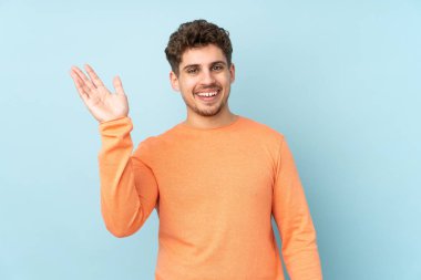 Beyaz adam mavi arka planda izole edilmiş, mutlu bir ifadeyle selamlıyor.