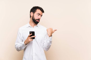 Sakallı genç adam, elinde cep telefonu var, mutsuz ve bir tarafı işaret ediyor.