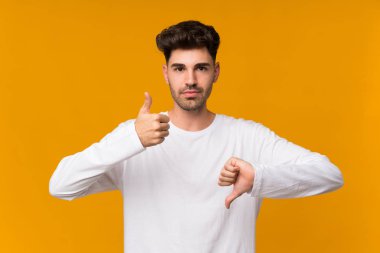 İzole edilmiş turuncu arka planı olan genç adam iyiye-kötüye işaret yapıyor. Evet ya da hayır arasında kararsız