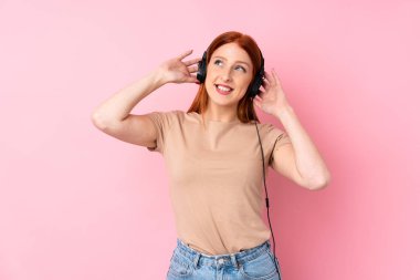 Genç kızıl saçlı kadın pembe arka planda kulaklıklı cep telefonunu kullanıyor.