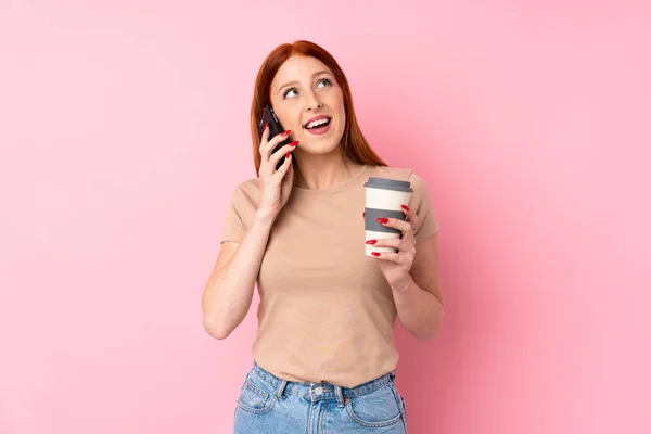 Giovane Donna Rossa Sfondo Rosa Isolato Con Caffè Portare Cellulare — Foto Stock