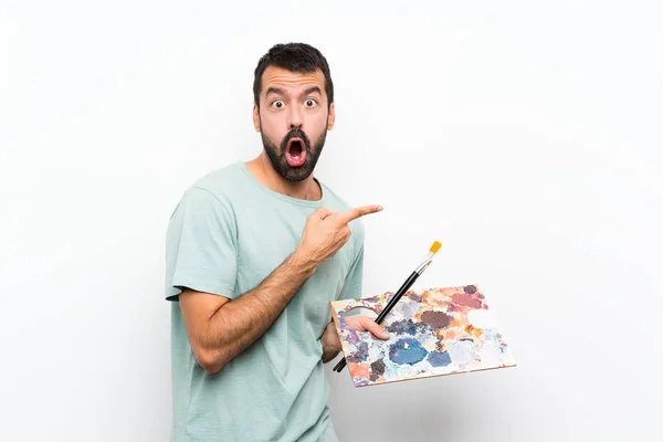 年轻的艺术家男人拿着调色板在孤立的背景下惊讶地指向一边 — 图库照片