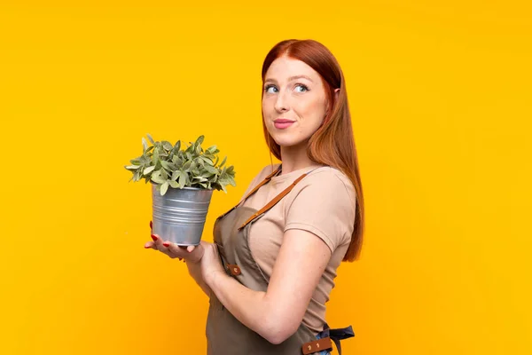 Junge Rothaarige Gärtnerin Hält Eine Pflanze Vor Isoliertem Gelben Hintergrund — Stockfoto