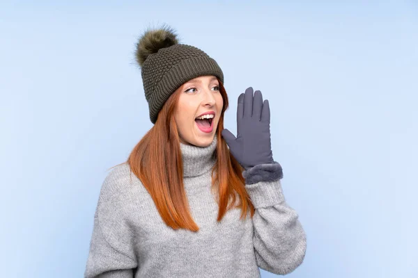 Young redhead Russian woman with winter hat over isolated blue background shouting with mouth wide open