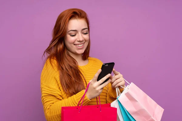 赤毛ティーン女の子上の隔離された紫色の背景保持ショッピングバッグと書き込みメッセージとともに彼女の携帯電話に友人 — ストック写真
