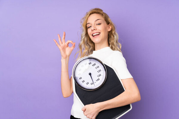Young blonde woman isolated on purple background holding a weighing machine and doing OK sign