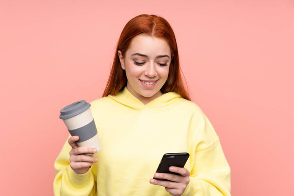 Redhead teenager girl over isolated pink background holding coffee to take away and a mobile