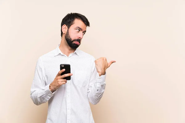 Joven Con Barba Sosteniendo Móvil Infeliz Señalando Hacia Lado —  Fotos de Stock