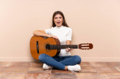 Mladá žena s kytarou sedí na podlaze s překvapivým výrazem ve tváři