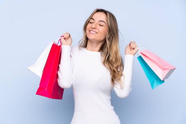 Soyutlanmış mavi arka planı olan genç bir kadın alışveriş torbaları tutuyor ve gülümsüyor.