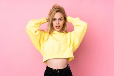 Pembe arka planda izole edilmiş sweatshirt giyen genç sarışın kadın hayal kırıklığına uğradı ve ellerini kafasından tuttu.
